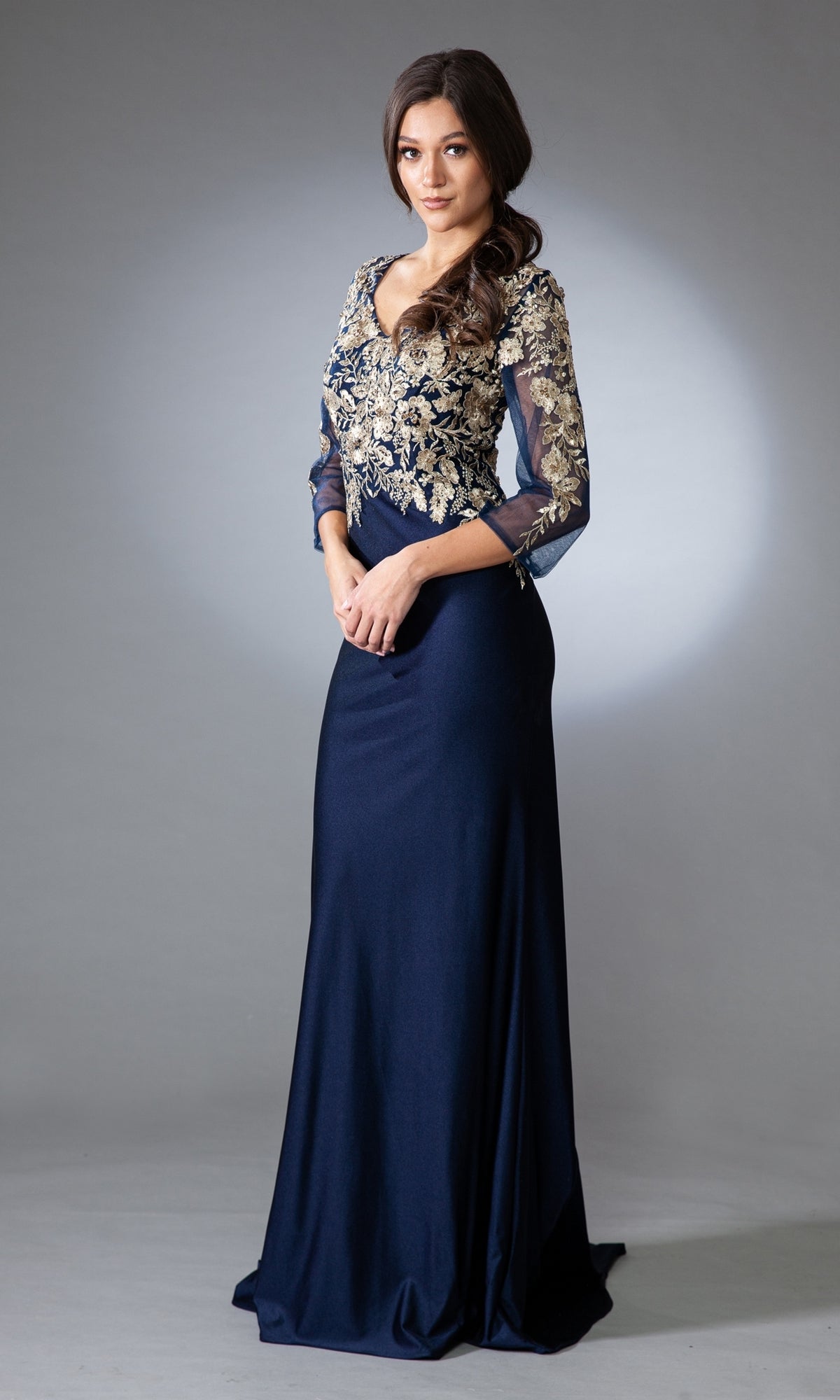 A woman wears the Amelia Couture 7039 navy blue mother-of-the-bride dress, featuring an embroidered bodice and sheer long sleeves, standing elegantly against a plain gray background.